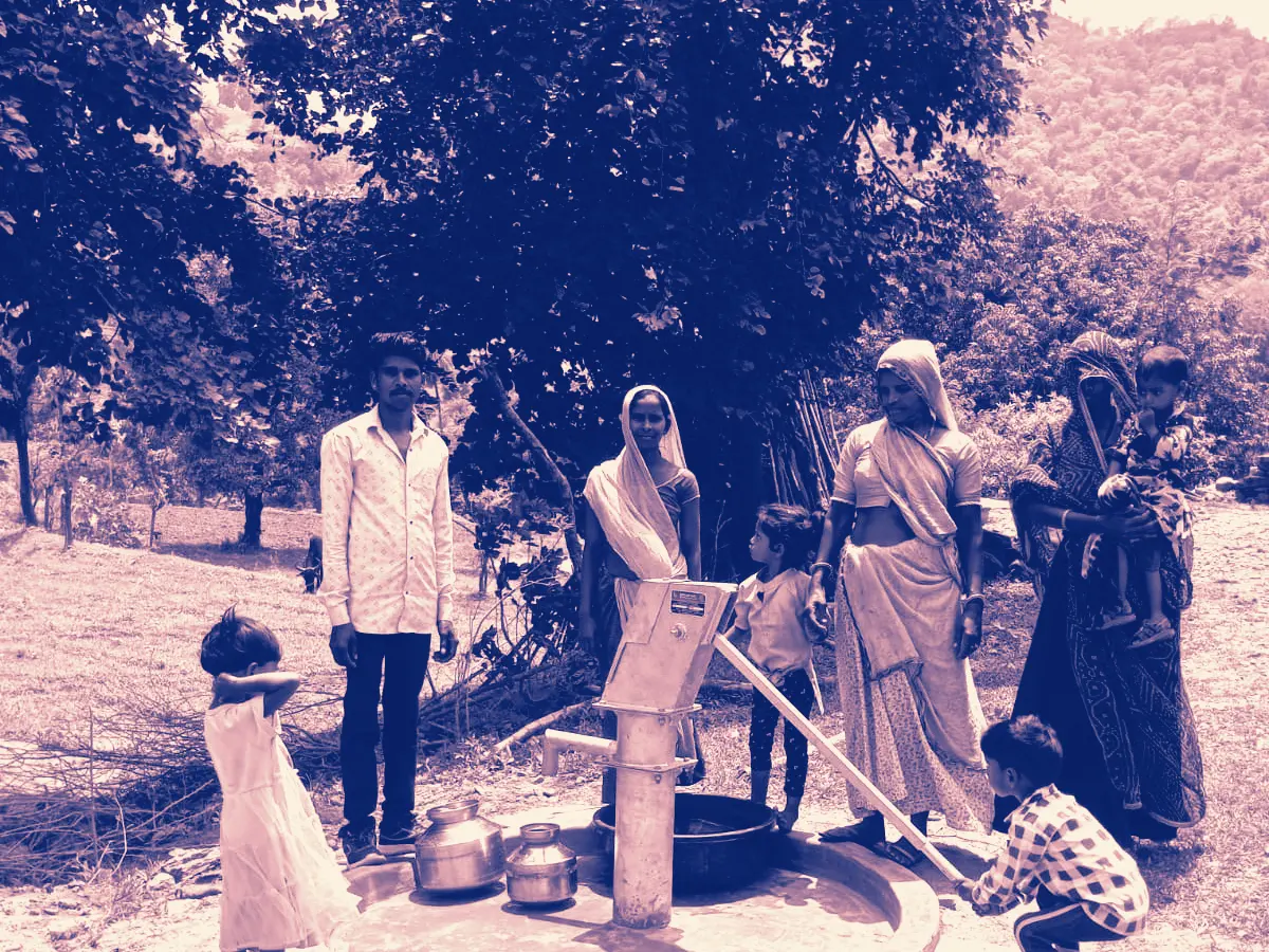 Two women drawing water at a hand pump on the edge of a village field